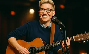 A person with short blond hair, glasses, and a blue sweater plays an acoustic guitar and sings into a microphone.