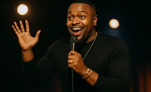 A Black man smiles and waves while holding a microphone on stage.