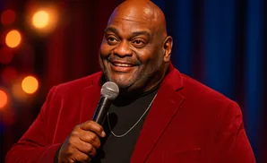 A bald Black man with a gray beard smiles and holds a microphone while wearing a dark suit and light blue shirt.
