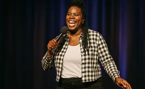 A Black woman with braided hair laughs while holding a microphone on a stage with a dark blue curtain background.