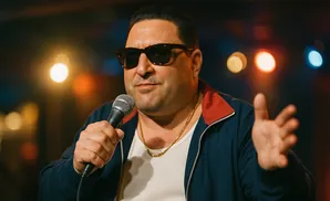 A man in sunglasses and a blue jacket with a red collar speaks into a microphone.