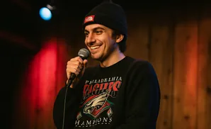 A smiling man in a beanie and sweatshirt holds a microphone on stage.