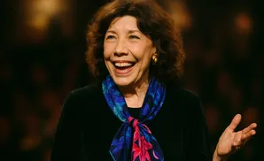 A woman with curly brown hair and a bright smile wears a black top and a blue and pink floral scarf.