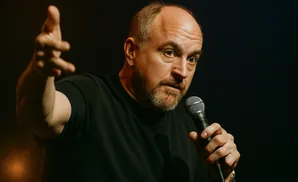 Comedian Louis C.K. speaking into a microphone with his left hand raised.