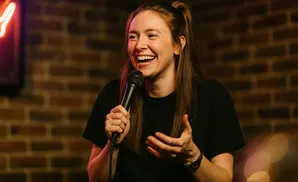 A woman with her hair in a ponytail laughs while holding a microphone, with a brick wall in the background.
