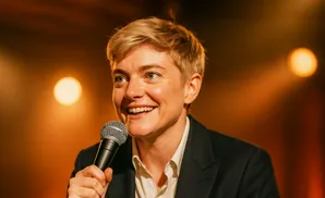 A smiling person with short blonde hair holds a microphone and wears a dark suit with a white shirt.