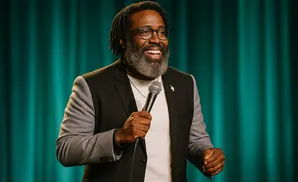 A Black man with glasses and a gray beard smiles holding a microphone.