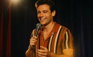 A man in a striped shirt holds a microphone and smiles at the camera.