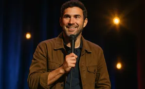 A smiling man in a brown jacket holds a microphone on a dark stage with blue curtains and warm lights.