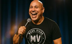 Bald man in a black t-shirt with a logo laughs while holding a microphone.