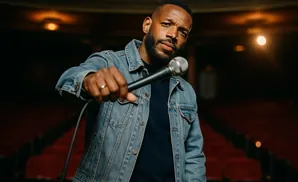 A Black man in a denim jacket holds a microphone toward the camera in a theater with red seats.