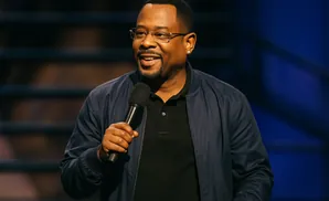 Martin Lawrence smiles and holds a microphone onstage, wearing a dark jacket over a black shirt.