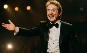 A man in a tuxedo and bow tie smiles widely with his arm outstretched, as if addressing an audience.