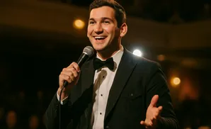 A man in a tuxedo and bow tie smiles and holds a microphone, speaking into it.