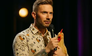 A man in a chicken-print shirt holds a microphone and a chicken puppet while speaking.