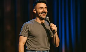 A man with a mustache smiles and holds a microphone onstage in front of blue and brown curtains.
