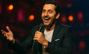 A man in a suit smiles broadly while holding a microphone and gesturing with his other hand on a dimly lit stage.