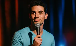 A man in a teal t-shirt holds a microphone and smiles at the camera on a dark stage with blue and orange lights.