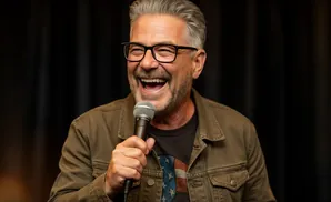 A man with gray hair and glasses laughs while holding a microphone.