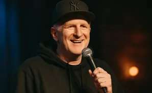 Comedian Michael Che, wearing a black Yankees hat and hoodie, smiles while holding a microphone.