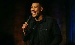 Comedian laughing into a microphone, wearing a denim jacket.