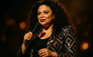 Woman with curly dark hair in a sparkly jacket holds a microphone and smiles.