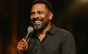 A Black man with a beard smiles widely into a microphone, wearing a dark collared shirt.