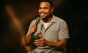 A Black man with a short haircut smiles as he holds a microphone and gestures with his hands.