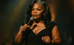 Mo'Nique speaks into a microphone on stage, wearing a black dress.