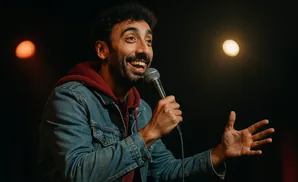 A man in a denim jacket and maroon hoodie smiles while holding a microphone and gesturing with his hands.