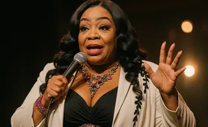 A Black woman speaks into a microphone, wearing a cream blazer, a black top, and a colorful jeweled necklace and bracelet.