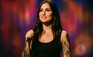 A woman with long dark hair and tattoos holds a microphone, smiling with a colorful blurred background.