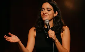 A woman with glasses and dark hair holds a microphone and gestures with her left hand while speaking.
