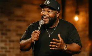 Comedian wearing a black LA Dodgers hat and shirt, holding a microphone and laughing.