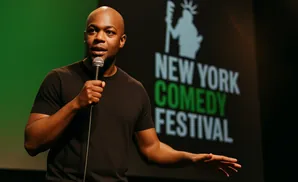 A bald Black man in a black t-shirt holds a microphone on stage, speaking in front of a 
