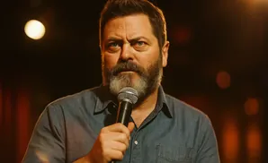 Nick Offerman holds a microphone, looking seriously at the camera in front of a dark, blurred background.
