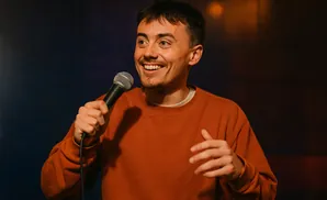 A man in an orange sweatshirt holds a microphone and smiles, gesturing with his hands.