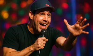 A comedian in a black t-shirt and baseball cap holds a microphone, animatedly speaking into it with his hand raised.