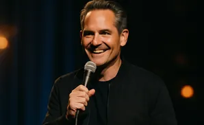 A man with graying hair smiles widely, holding a microphone in his right hand. He wears a black bomber jacket.