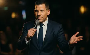 Man in a suit speaks into a microphone, gesturing with his other hand.