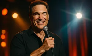 A smiling man in a black shirt holds a microphone on a dimly lit stage with warm orange and bright white spotlights.