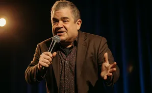 Comedian Patton Oswalt holds a microphone and gestures with his hand while on stage.