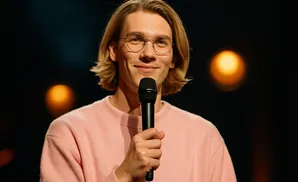 A smiling man with blond hair and glasses holds a microphone.