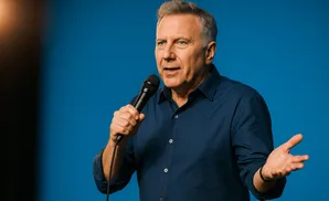 A man in a blue shirt holds a microphone and gestures with his other hand against a blue background.