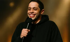 Pete Davidson smiles while holding a microphone and wearing a black hoodie.