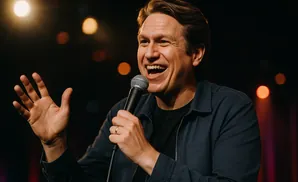 Comedian Pete Davidson on stage, laughing with a microphone in his hand and his other hand raised.