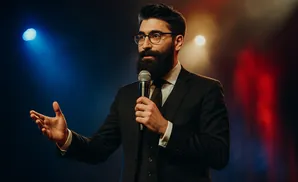 Man in suit with beard and glasses speaking into a microphone on stage with blue and red lighting.