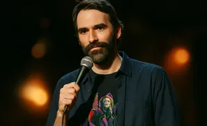 A man with a beard stands with a microphone, wearing a dark blue shirt over a graphic t-shirt.