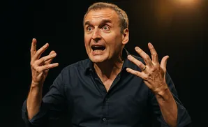 A man in a dark blue shirt performs at a microphone, animatedly gesturing with his hands.