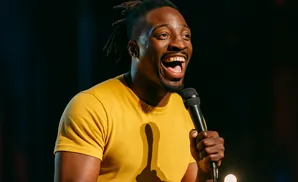 A Black man with dreadlocks laughs into a microphone while wearing a yellow t-shirt.